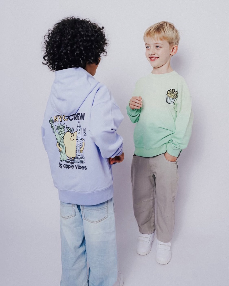 Two children in Fabro-made knitwear, one in a lilac hooded sweatshirt with a printed back graphic, one in a mint sweatshirt with an appliqu&eacute; chest motif